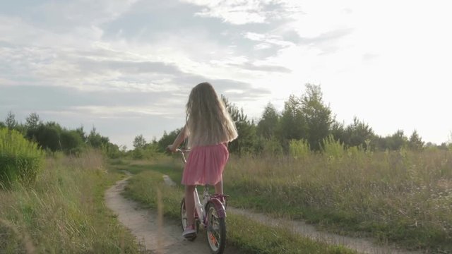 Happy, beautiful, little girl with long blond hair in a pink skirt and a jumper rides a kids bicycle on the road, she smiles. Sunny summer weather, the sun's rays