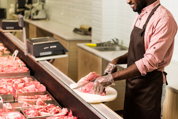 partial view of smiling african american male shop assistant in apron cutting steak of raw meat in...