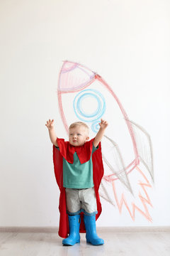 Adorable little child playing astronaut near wall with drawing of spaceship indoors