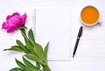 Empty notepad with place for text, pen, a cup of tea and peony flower on a white wooden background. Mockup for your design. View from above.