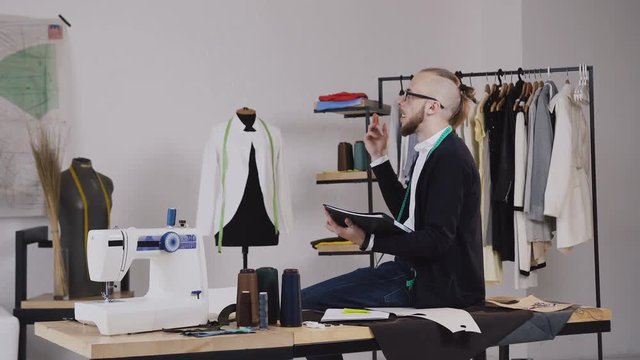 A handsome fashion designer sits on the table in the studio and draws sketches for tailoring the new collection of clothes. A young bearded tailor wearing glasses, dressed in a white shirt and a black