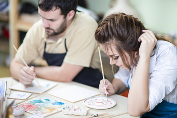 Two Young Caucasian Ceramists Painting and Glazing Clay Crafts Together in Workshop with Paintbrushes.