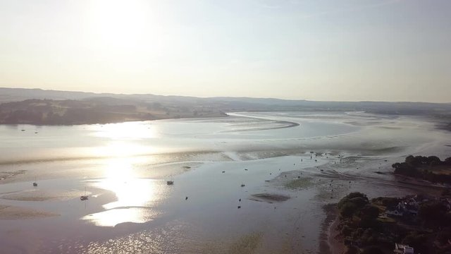 Drone gliding over the oceans water off of the shore of Lympstone England. The sun is reflecting in the water, and the boats are docked close to shore.