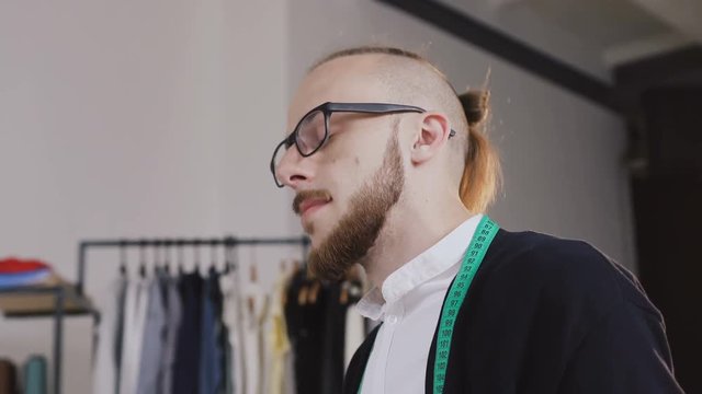 Close-up of a fashion designer who sits on the table in the atelier and draws sketches for tailoring the new collection of clothes. A bearded tailor wearing glasses, dressed in a white shirt and a
