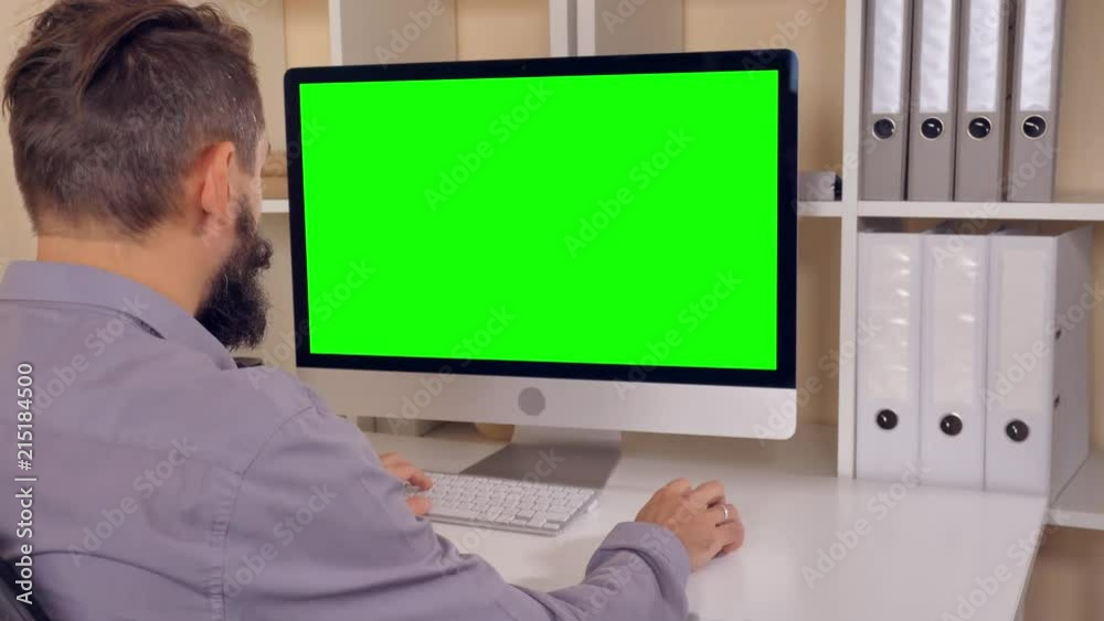 Poster man sitting at workplace paying game on computer in small office. businessman with beard looking on display with green screen. employee in startup company typing on keyboard use pc mouse