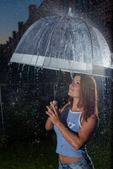 Woman in the rain with a transparent umbrella in the evening. Smiling young woman in the rain with umbrella in the evening. Beautiful woman with a transparent umbrella in the lanterns and rain drop