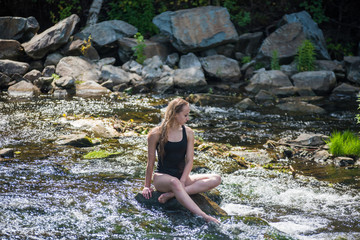 Young blond woman in a black swimwear sitting by the waterfall.  Young woman enjoying nature