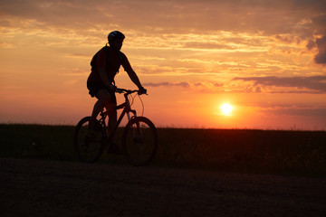 Fototapeta na wymiar Silhouette of a bicyclist traveling on a sunset background on the road to the field