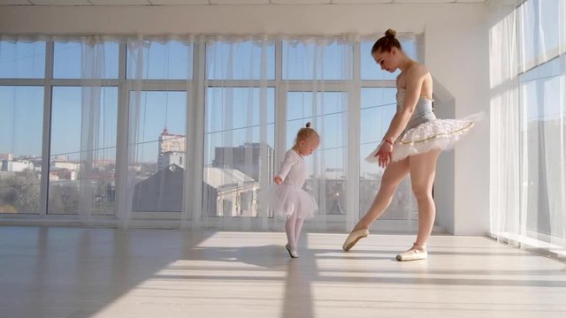 Young gorgeous ballerina teaches daughter dancing in studio. Happy ballerina with her child girl standing on tiptoes. Mother and daughter dancing ballet