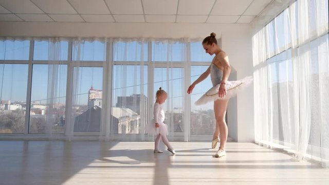 Young gorgeous ballerina teaches her little daughter dancing in studio. Happy ballerina with her child girl standing on tiptoes. Mother and daughter dancing ballet