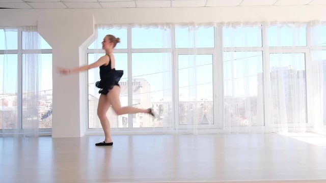 Graceful young ballerina in black bodysuit jumping in bright white studio. Young dancer performing a jump in the air