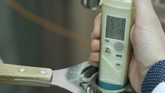 Close-up of hand of brewery worker holding digital refractometer into steel cylinder with beer
