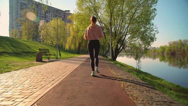 female running in the early morning outdoors. back view woman wearing black leggings and pink hoodie jogging along the river in spring season