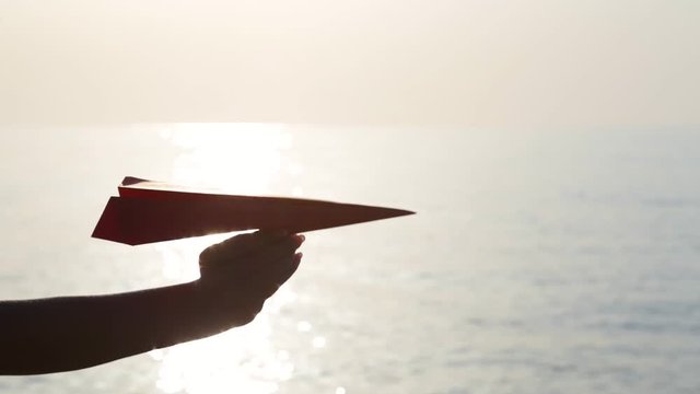 Closeup view of paper red airplane in hand of young woman isolated on sunny sunset sky and sea water background.  Dreaming about travelling concept. Video shoot at golden hour on summer morning.
