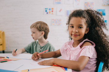 selective focus of multicultural preschoolers at table with papers and pencils in classroom