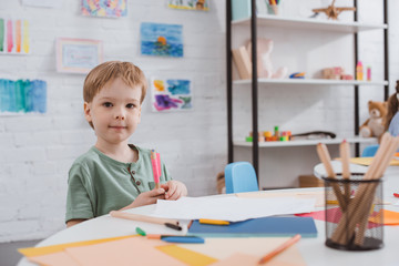 Fototapeta premium portrait of preschooler boy sitting at table with paper and colorful pencils for drawing in classroom