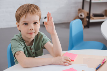 portrait of cute preschooler with hand up sitting at table in classroom
