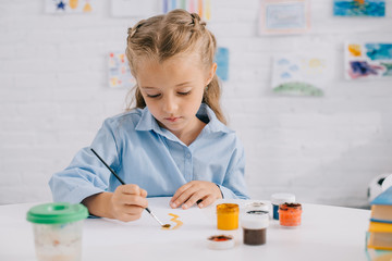 portrait of adorable focused child drawing picture with paints and brush at table