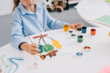 partial view of child drawing picture with paints and paint brush at table