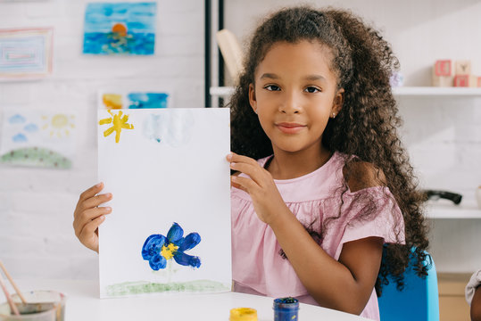 portrait of african american kid showing colorful picture in hands while sitting at table in room