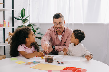 smiling caucasian teacher in eyeglasses and african american kids drawing pictures with paints together in classroom