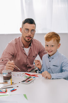 portrait of teacher and little boy with paint brushes sitting at table in classroom