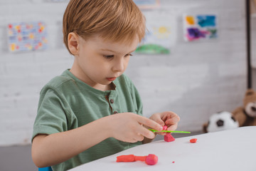 focused boy with colorful plasticine sculpturing figure at table in room