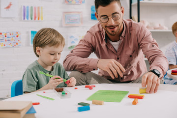 portrait of teacher and adorable preschooler with plasticine sculpturing figures at table in classroom - obrazy, fototapety, plakaty