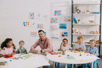smiling teacher and multiethnic kids sitting at table with colorful plasticine in classroom