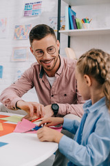 smiling teacher and cute preschooler cutting papers with scissors att able in classroom
