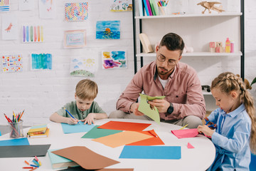 portrait of focused teacher and preschoolers cutting colorful papers with scissors in classroom