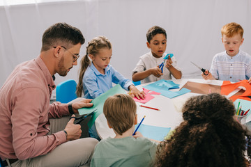 partial view of male teacher and multiracial preschoolers cutting colorful papers with scissors at...