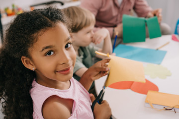 selective focus of multiethnic preschoolers cutting colorful papers with scissors in classroom