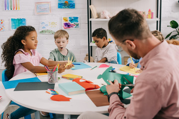 teacher and multiracial preschoolers with colorful papers and scissors making paper applique in...
