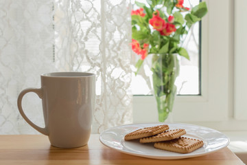 Morning time. White cup of coffee or tea, white plate with cookieon a wooden table. Vase with flowers on the window. Breakfast, lunch