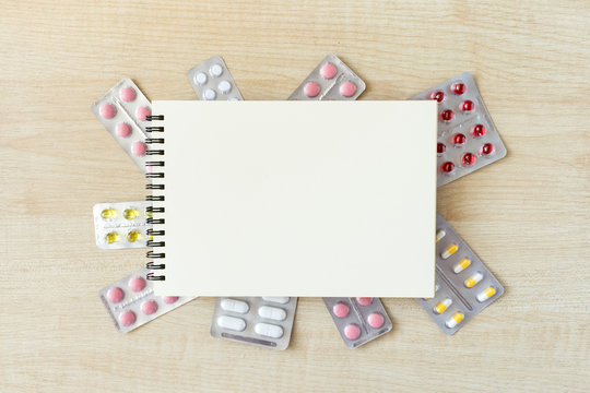 Paper notepad mock up in frame of various blisters with multi-colored tablets and capsules on wooden table. Doctor desk background, horizontal medical concept copy space, white mockup