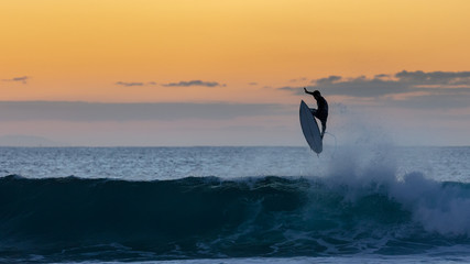 The silhouette of a surfer in the air above a wave at sunrise with a golden sky in the background - Powered by Adobe