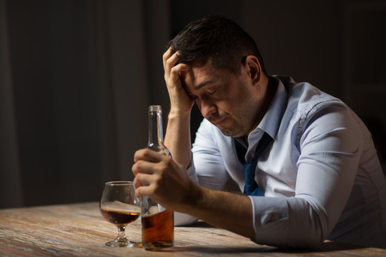 alcoholism, alcohol addiction and people concept - male alcoholic with bottle drinking brandy at table at night