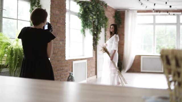 Backside view of a photoshooting process. Beautiful young pregnant photo model in white peignoir posing for female photographer in modern decorated room-studio