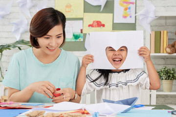 Little Asian girl sitting at teacher and having fun with paper with cut-out heart in art class
