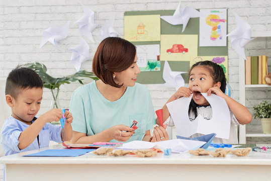 Little Asian girl looking at teacher and having fun with paper with cut-out heart