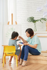 Asian teacher woman helping to girl with painting at easel in art class