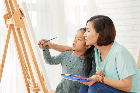 Side view of Asian woman teaching little girl to draw at easel in art class