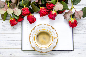Red roses, diary, coffee and saucer on a light wooden table, flat lay