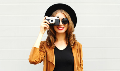 Cool funny girl model with retro film camera wearing a elegant hat, brown jacket, curly hair outdoors over city grey background