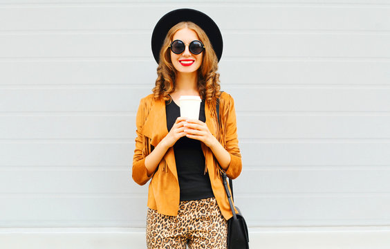 Elegant woman with coffee cup wearing retro elegant hat, sunglasses, brown jacket and black handbag over grey background
