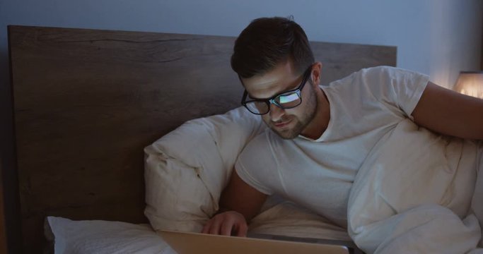 Portrait of the handsome young caucasian man lying in the bed in the evening in his glasses and chatting or scrolling on the laptop computer.
