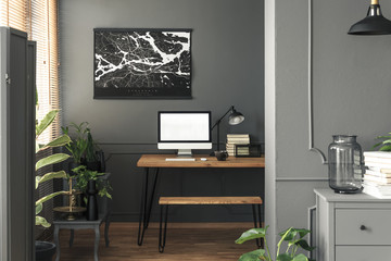 Real photo of dark open space living room interior with map poster on the wall above wooden desk for remote work with mockup computer, books and lamp
