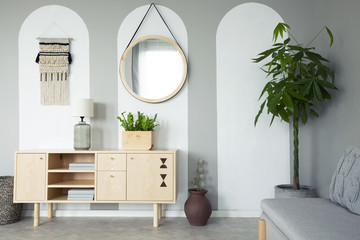 Round mirror hanging on the wall in real photo of grey living room interior with retro cupboard...