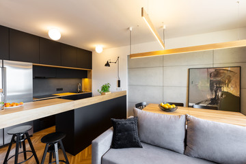 Lights above countertop in open black kitchen interior with grey sofa and dining table. Real photo